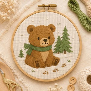 May include: A circular wooden embroidery hoop containing a fabric panel with a stitched design of a brown bear wearing a green scarf. The bear sits in a snowy landscape with green pine trees and small paw prints embroidered on the white fabric.