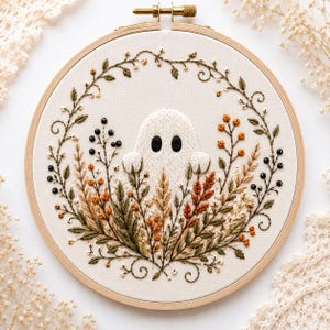 May include: A circular wooden embroidery hoop containing a fabric panel with a hand-stitched design of a white ghost surrounded by a wreath of embroidered autumn-coloured wheat, leaves, and berries in shades of orange, brown, and dark green.