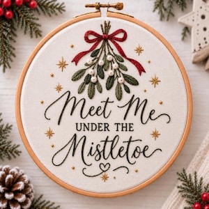 Könnte beinhalten: Ein runder hölzerner Stickrahmen mit weißem Stoff, der ein handgesticktes Mistelzweig-Motiv mit roter Schleife zeigt. Der Text "Meet Me UNDER THE MISTLETOE" ist in schwarzer Schrift gestickt und wird von kleinen goldenen Sternen und Punkten umrahmt.
