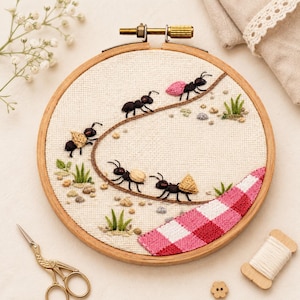 May include: A circular wooden embroidery hoop containing a piece of off-white fabric with a hand-stitched design of five black ants carrying various small items along a curved brown line, with a pink and white chequered picnic blanket corner.