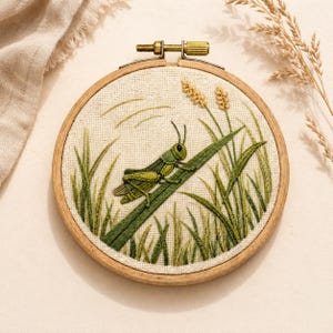 Könnte beinhalten: Ein runder hölzerner Stickrahmen mit einem cremefarbenen Stoff, der ein handgesticktes Motiv einer grünen Heuschrecke auf einem langen Grashalm zeigt, umgeben von verschiedenen in Grüntönen gestickten Grashalmen.