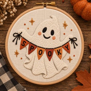 May include: A circular wooden embroidery hoop containing a cream-coloured fabric with a hand-stitched ghost design. The ghost is white with black eyes, a smile, and pink cheeks, holding a banner of orange and brown pennants that spell SPOOKY.