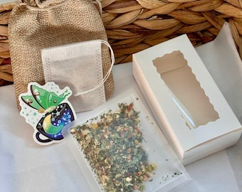 May include: A small hessian bag with a drawstring closure, containing a tea bag. The tea bag is filled with loose leaf tea and is sitting on a white surface. A sticker of a teacup with a butterfly and stars is next to the bag. A white box with a window is also in the image.