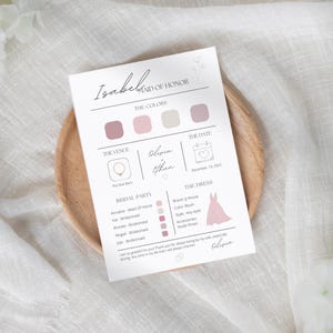 May include: A white invitation card with the text "Isabel Maid of Honor" and details about a wedding. The card includes color swatches, venue, date, bridal party, and dress information. The card is on a wooden plate.