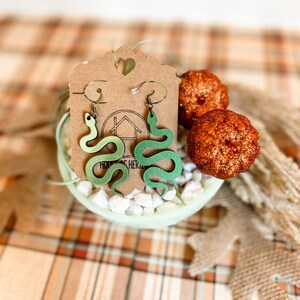 May include: A pair of green snake earrings with a gradient effect, displayed on a small, light green dish filled with white stones. The earrings hang from a brown tag with a heart-shaped cutout. Two glittery orange decorative pumpkins are also in the image.