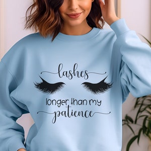 May include: A light blue sweatshirt with the text "lashes longer than my patience" in black script. The design includes a pair of stylized black eyelashes above the text. The sweatshirt is a solid color and has long sleeves.