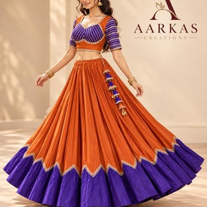 May include: A two-piece lehenga set featuring a vibrant orange and deep purple color scheme. The blouse has short sleeves with purple and orange stripes, while the long, pleated skirt includes a wide purple hem with a zigzag gold trim.