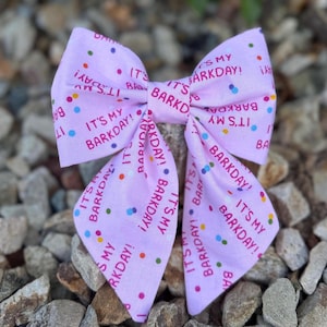 May include: A light pink dog bow tie with the text "IT'S MY BARKDAY!" and colorful confetti dots. The bow tie is made of fabric and is designed to be worn by a dog.