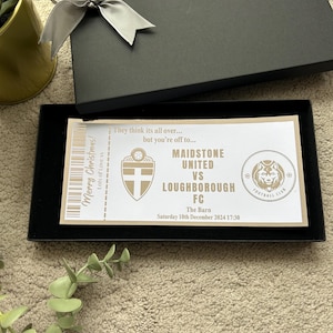 May include: A black gift box with a silver bow, containing a decorative ticket. The ticket reads "Maidstone United vs Loughborough FC" with the date "Saturday 10th December 2024 17:30". The text "Merry Christmas! Lots of Love xx" is also visible.