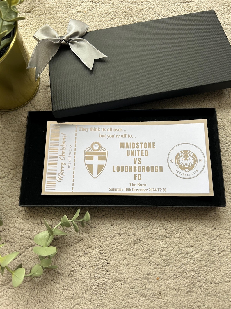 May include: A black gift box with a silver bow, containing a decorative ticket. The ticket reads "Maidstone United vs Loughborough FC" with the date "Saturday 10th December 2024 17:30". The text "Merry Christmas! Lots of Love xx" is also visible.
