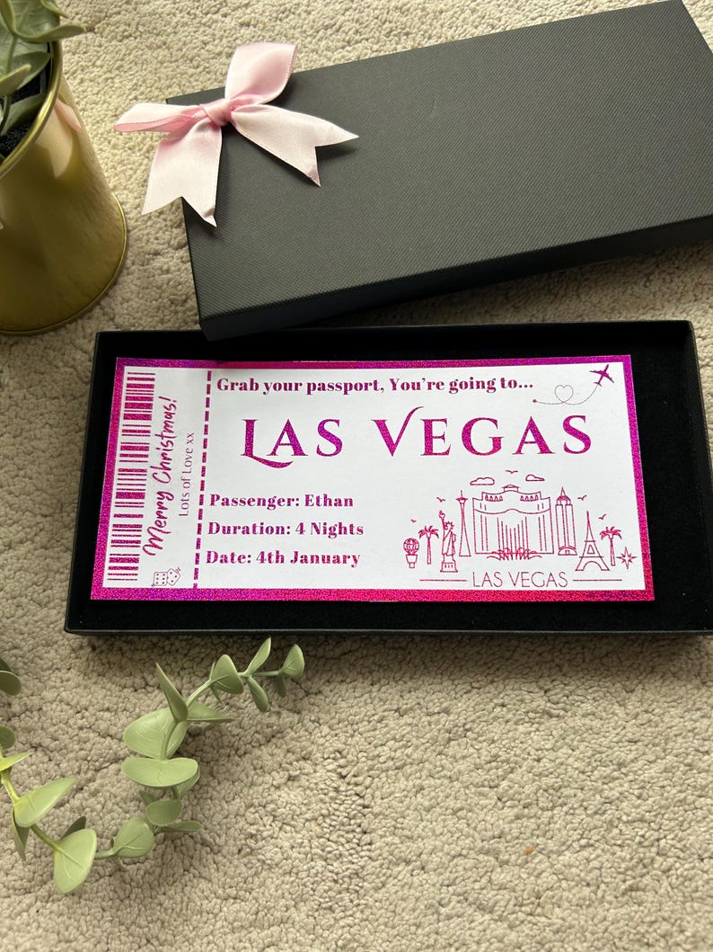 May include: A gift box with a pink bow on top, containing a Las Vegas ticket. The ticket has the text "Merry Christmas!" and "Las Vegas" in pink, along with details like passenger name, duration, and date.