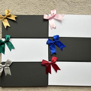 May include: Arrangement of six rectangular gift boxes, alternating between black and white. Each box is adorned with a satin bow in various colors: gold, pink, green, blue, silver, and red. The boxes are arranged on a neutral-toned surface.