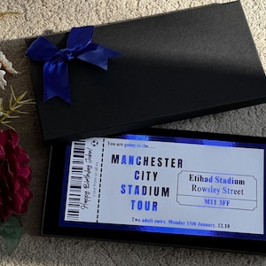 May include: A black gift box with a blue bow on top, open to reveal a ticket for a Manchester City Stadium Tour. The ticket has text that reads "Happy Birthday John!" and "Two adult entry. Monday 15th January. 12.10."