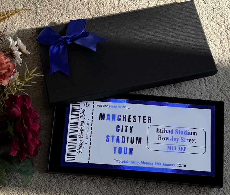 May include: A black gift box with a blue bow on top, open to reveal a ticket for a Manchester City Stadium Tour. The ticket has text that reads "Happy Birthday John!" and "Two adult entry. Monday 15th January. 12.10."