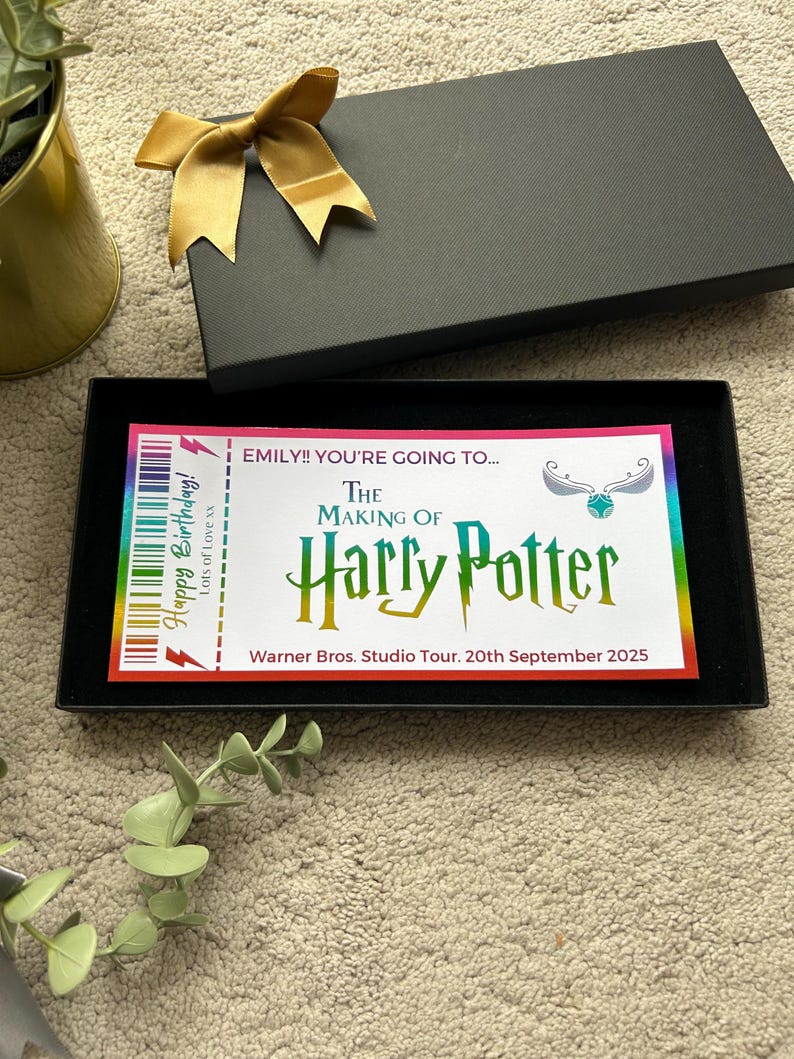 May include: A black box with a gold bow on top. The box is open and inside is a colorful ticket with the text "EMILY!! YOU'RE GOING TO... THE MAKING OF Harry Potter Warner Bros. Studio Tour. 20th September 2025".