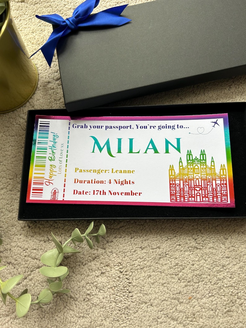 K&ouml;nnte beinhalten: Ein rechteckiges Geschenkticket mit Regenbogenrand in einer schwarzen Schachtel. Der Text lautet: Grab your passport, You're going to... MILAN, Happy Birthday! Lots of Love xx, Passenger: Leanne, Duration: 4 Nights, Date: 17th November. Enth&auml;lt eine Grafik einer Kathedrale.