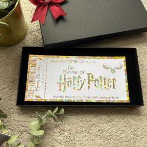 May include: A golden ticket with a holographic finish, reading "OLIVER!! YOU'RE GOING TO... THE MAKING OF HARRY POTTER Warner Bros. Studio Tour. 30th January 2025". The ticket is in a black box with a red bow.