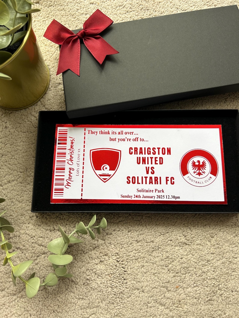 May include: A black gift box with a red bow on top, containing a football ticket. The ticket is red and white, with the text "Merry Christmas!" and details for a match between Craigston United and Solitari FC.