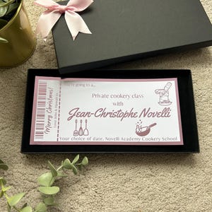 K&ouml;nnte beinhalten: Eine schwarze rechteckige Geschenkbox mit einer hellrosa Schleife auf dem Deckel. Im Inneren befindet sich ein rechteckiger Gutschein mit dem Text: Merry Christmas! Love John & Hannah xx, You're going to a... Private cookery class with Jean-Christophe Novelli.