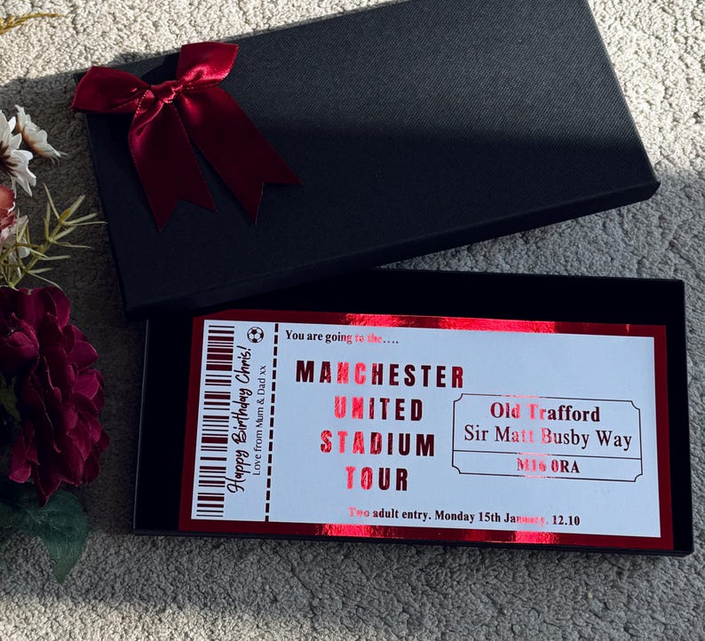 May include: A black gift box with a red bow, open to reveal a Manchester United Stadium Tour ticket. The ticket is red and white with text that reads "Happy Birthday Chris!" and "Love from Mum & Dad xx".