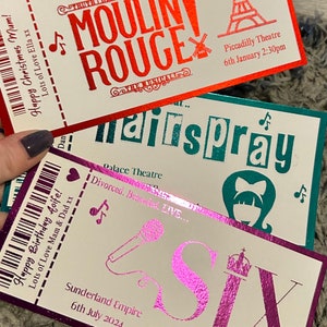 K&ouml;nnte beinhalten: Drei rechteckige Theatertickets mit metallischer Folienbeschriftung und Grafiken. Das oberste Ticket ist rot f&uuml;r Moulin Rouge, das mittlere ist blaugr&uuml;n f&uuml;r Hairspray und das untere ist lila f&uuml;r Six. Jedes Ticket enth&auml;lt einen Strichcode, Noten und Veranstaltungsdetails.