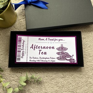 K&ouml;nnte beinhalten: Eine rechteckige schwarze Geschenkbox mit einer blauen Schleife, die ein wei&szlig;es Ticket mit violettem Text und Grafiken enth&auml;lt. Das Ticket lautet: Mum, A Treat for you... Afternoon Tea, Happy Birthday! The Rubens, Buckingham Palace, 14. Februar 12.30 Uhr.