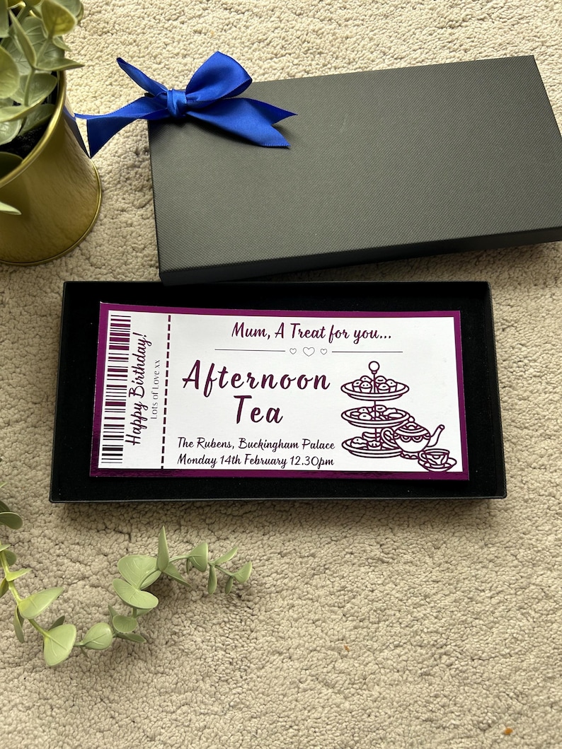 K&ouml;nnte beinhalten: Eine rechteckige schwarze Geschenkbox mit einer blauen Schleife, die ein wei&szlig;es Ticket mit violettem Text und Grafiken enth&auml;lt. Das Ticket lautet: Mum, A Treat for you... Afternoon Tea, Happy Birthday! The Rubens, Buckingham Palace, 14. Februar 12.30 Uhr.