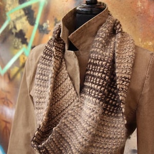 Könnte beinhalten: Ein brauner und schwarzer gestrickter Infinity-Schal mit einem strukturierten Muster. Der Schal wird über einer braunen Jacke getragen.