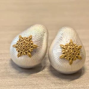 May include: A pair of teardrop-shaped earrings featuring a brushed silver finish with fine vertical lines. Each earring is centered with a small, intricate gold-toned metal snowflake emblem that has a detailed, filigree-like floral pattern.