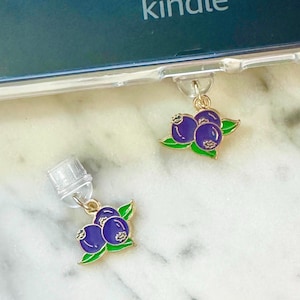 May include: Two small, gold-colored charms with a cluster of purple blueberries and green leaves. The charms are attached to a clear plastic case, with the word "kindle" visible on the device.