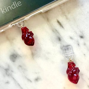 May include: Clear phone case with two red heart-shaped charms. The charms are attached to the case with small gold-colored hooks. The word "kindle" is visible on the top of the phone case.