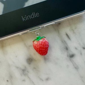 May include: A red strawberry charm with green leaves hangs from a Kindle e-reader. The e-reader is black with the word "kindle" printed in white. The background is a white and gray marble surface.