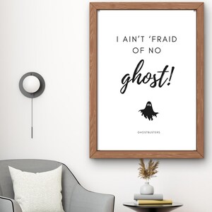 May include: Printable digital download of a black and white illustration of a ghost with the text "I ain't 'fraid of no ghost!" and "Ghostbusters" below the ghost. The illustration is framed in a brown wood frame and is hanging on a white wall.