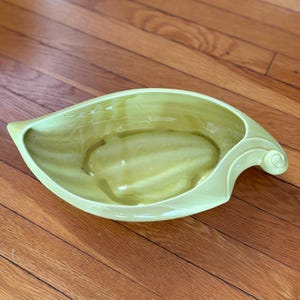 Può includere: Un vaso da fiori in ceramica verde chiaro a forma di foglia con un manico curvo. Il vaso ha una finitura liscia e lucida.