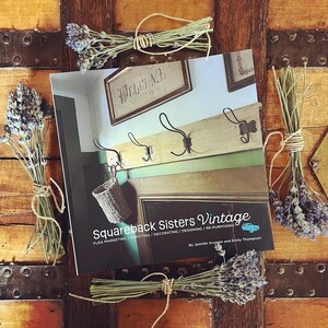 May include: A book titled "Squareback Sisters Vintage" with the subtitle "Flea Marketing/Thrifting/Decorating/Designing/Re-Purposing" by Jennifer Krueger and Emily Thompson. The book cover features a vintage interior with a wooden coat rack and a welcome sign.