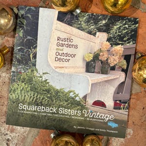 May include: A book cover with a white wooden bench and flowers in front of a green leafy background. The title is "Rustic Gardens and Outdoor Decor" and the subtitle is "Squareback Sisters Vintage Flea Marketing/Thrifting/Decorating/Designing/Re-Purposing" by Jennifer Krueger and Emily Thompson.