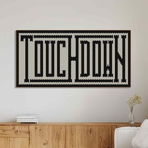 May include: A black and white wall art piece with the word "TOUCHDOWN" in a bold, geometric font. The artwork is framed in black and hangs above a wooden cabinet and white sofa, creating a modern, minimalist aesthetic.