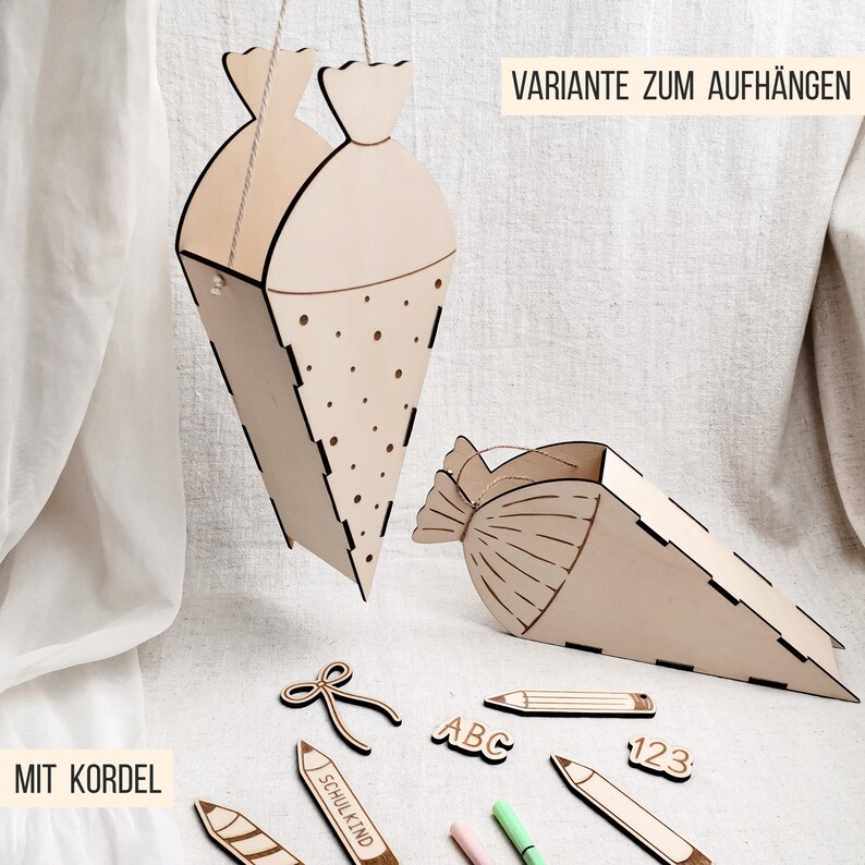 Puede incluir: Decoraciones de cono escolar de madera, una colgante y otra tumbada, con recortes decorativos. Incluye recortes de madera de l&aacute;pices, un lazo y las letras "ABC" y los n&uacute;meros "123". El texto "MIT KORDEL" y "VARIANTE ZUM AUFH&Auml;NGEN" son visibles.