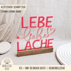 Puede incluir: Una base de madera sostiene un letrero decorativo rosa y blanco. El letrero presenta las palabras alemanas "LEBE LIEBE LACHE" en rosa, con un "Liebe" en cursiva en blanco. El letrero mide 16 cm de largo.