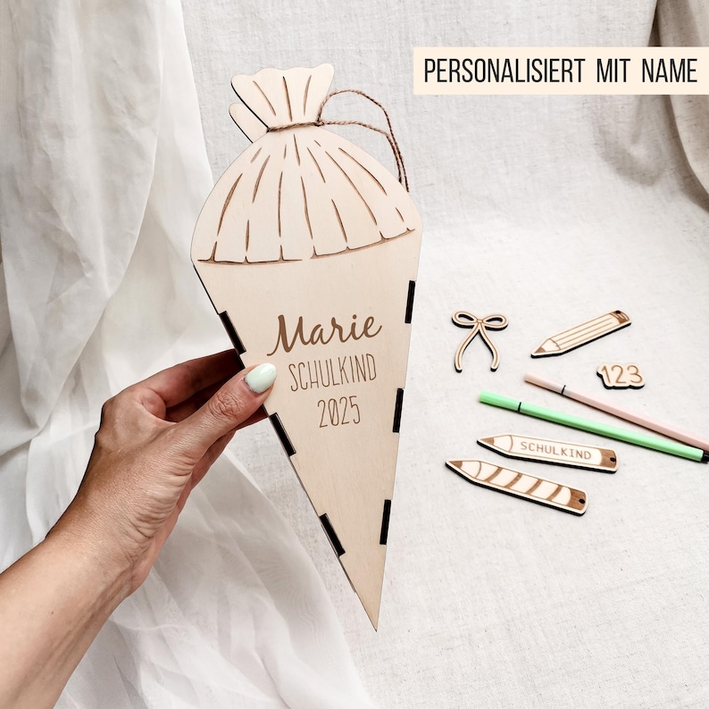 Puede incluir: Un cono escolar de madera con el nombre "Marie", la palabra "SCHULKIND" y el a&ntilde;o "2025" grabados. Acompa&ntilde;ado de recortes de madera de &uacute;tiles escolares, incluyendo l&aacute;pices y tijeras. Tambi&eacute;n se ve el texto "PERSONALISIERT MIT NAME".