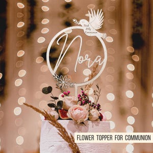 Pode incluir: Topper de bolo de madeira branco com um c&iacute;rculo, o nome "Nora" escrito no interior, e uma pomba com o texto "Primeira Comunh&atilde;o". O topper est&aacute; colocado em um bolo branco com flores rosa e roxas.