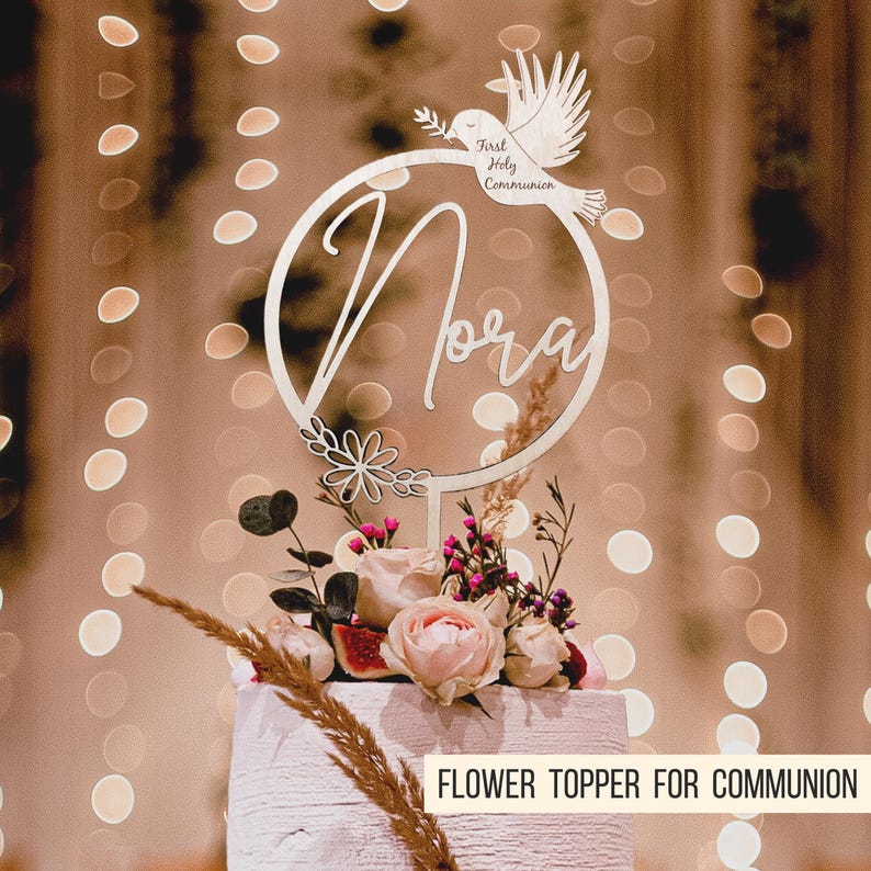 Pode incluir: Topper de bolo de madeira branco com um c&iacute;rculo, o nome "Nora" escrito no interior, e uma pomba com o texto "Primeira Comunh&atilde;o". O topper est&aacute; colocado em um bolo branco com flores rosa e roxas.