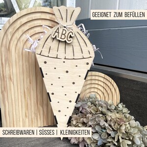 Puede incluir: Una bolsa escolar de madera en forma de cono, con las letras "ABC". La bolsa est&aacute; hecha de madera clara y tiene un dise&ntilde;o decorativo. El texto "GEEIGNET ZUM BEF&Uuml;LLEN" es visible. La parte inferior de la imagen tiene el texto "SCHREIBWAREN | S&Uuml;SSES | KLEINIGKEITEN".