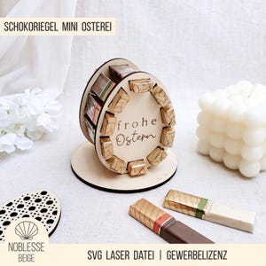 Può includere: Espositore in legno a forma di uovo di Pasqua con la scritta "Frohe Ostern". L'espositore contiene piccole barrette di cioccolato. Le parole "SCHOKORIEGEL MINI OSTEREI" sono in alto. Due barrette di cioccolato sono in primo piano.