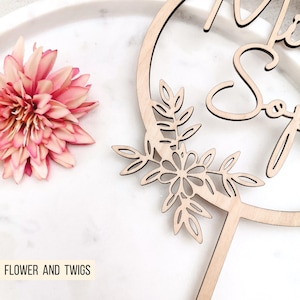 Pode incluir: Topper de bolo de madeira com um design circular e as palavras "Mr & Mrs" em it&aacute;lico. O c&iacute;rculo &eacute; decorado com um design floral com folhas e flores.