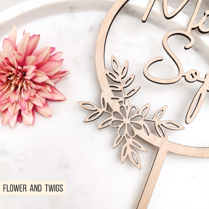 Pode incluir: Topper de bolo de madeira com um design circular e as palavras "Mr & Mrs" em it&aacute;lico. O c&iacute;rculo &eacute; decorado com um design floral com folhas e flores.