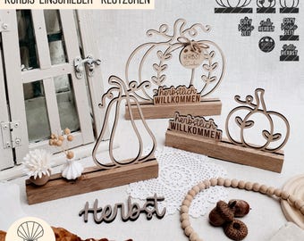 May include: Wooden fall-themed decorations featuring pumpkin and pear shapes. The decorations include the word "Herbst" and "herbstlich WILLKOMMEN". The items are displayed on a white surface with a window in the background.