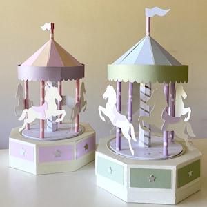 May include: Two paper carousel models featuring pastel-colored roofs, central poles, and white horse figures. The left carousel has a pink and yellow roof with a cream base, while the right carousel has a blue and green roof with a cream base.
