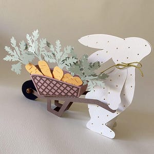 Puede incluir: Un conejito de papel blanco con lunares dorados empuja una carretilla marrón llena de zanahorias naranjas y follaje verde. La carretilla tiene una cesta rosa. Una cuerda dorada está atada alrededor del cuello del conejo.