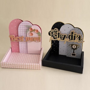 Puede incluir: Dos piezas decorativas de exhibición. Una tiene patrones de cuadros y lunares rosas y blancos, con "Best mom" en letras doradas. La otra tiene patrones negros, blancos y de cuadros, con "Feliz dia" en letras doradas y un trofeo.